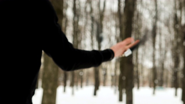 一个活跃的年轻人的手的特写，一个格斗家，在黑色的衣服，表演双截棍的技术在冬天，覆盖着雪的城市公园在树木之间视频下载