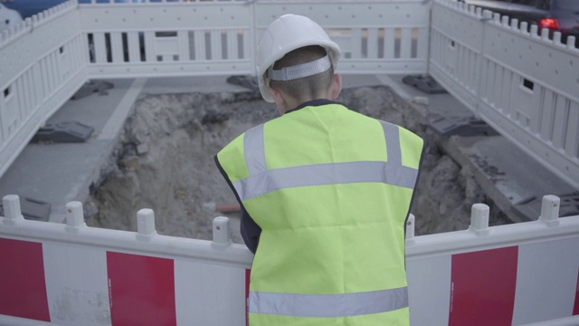 自信的小男孩穿着安全装备和建筑工人头盔站在道路上，检查坑道上。视频素材