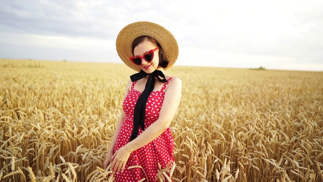 麦田里时髦女人的肖像。传统女孩草帽，心形眼镜，红色圆点裙子和口红。旅游、时尚、自然理念视频素材