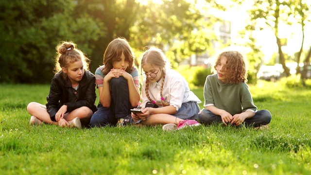 孩子和小玩意。同学们坐在公园的草地上互递智能手机。日落视频下载