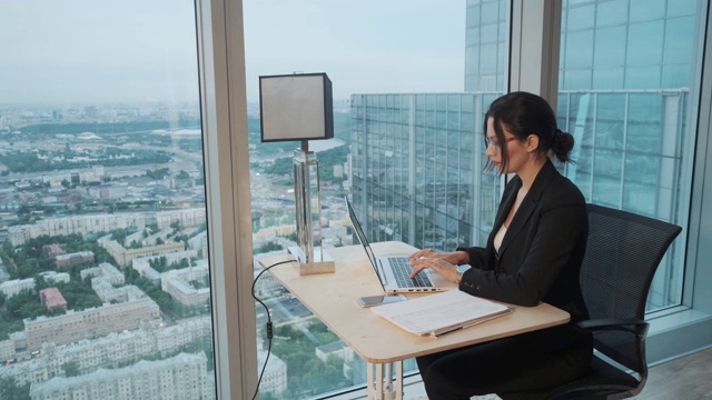商务女性在位于摩天大楼高层的现代办公室工作。漂亮的女孩坐在大窗户旁边的桌子上用笔记本电脑工作。视频下载