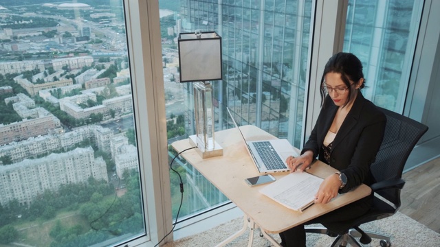 在摩天大楼高层的一间现代办公室里，年轻的商业女性使用笔记本电脑工作。视频下载