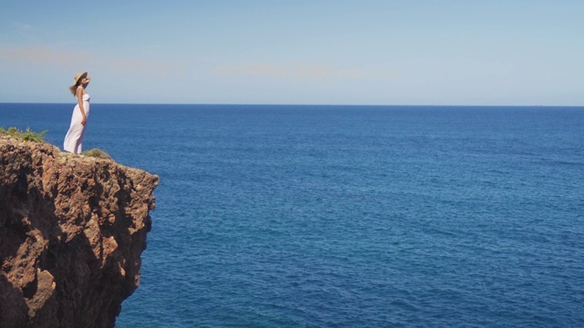 穿着白色连衣裙的女孩站在悬崖峭壁上。一个女人望着远处无边无际的海面视频素材