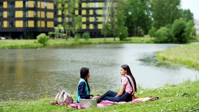 两个大学女生坐在公园池塘边的毯子上聊天。放松的学生，亚洲和高加索人，课后在自然中聊天，周围的书籍，笔记本电脑和背包视频下载