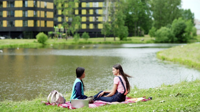 两个不同的女学生坐在公园池塘边的毯子上聊天。放松的大学朋友，亚洲和高加索人，喝咖啡和聊天在自然课后视频下载