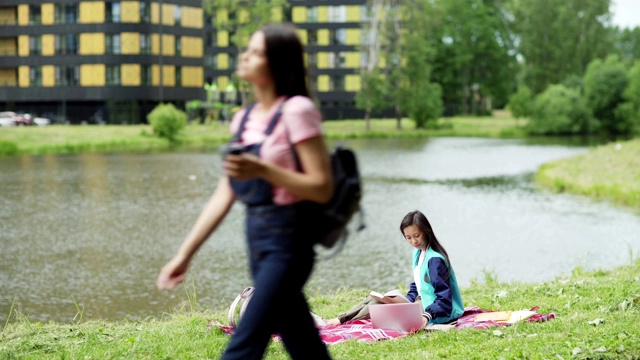 一个亚洲女孩坐在校园池塘边的毯子上，看书，用笔记本电脑打字。一个女学生在户外准备上课，另一个学生路过视频下载