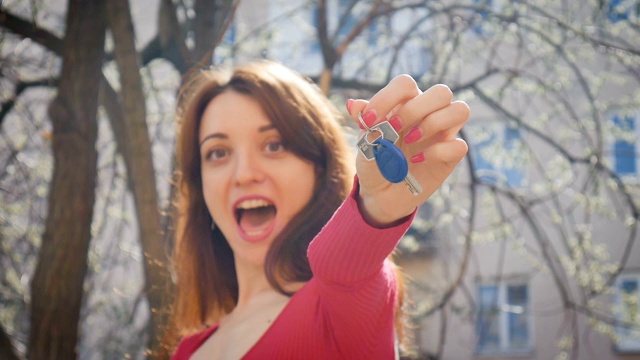情绪化的年轻女人拿着钥匙与蓝色的钥匙链与幸福和兴奋。女性的手与红色美甲握钥匙在户外在春天的城市背景。视频素材