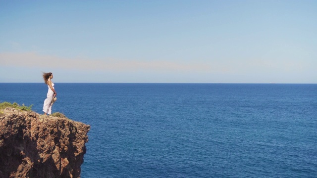穿着白色连衣裙的女孩站在悬崖峭壁上。一个女人望着远处无边无际的海面视频素材
