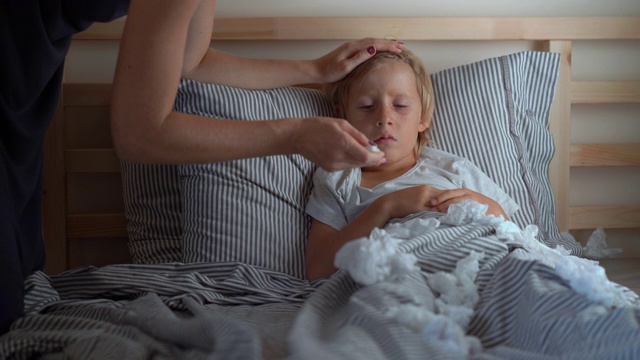 一个生病的小男孩躺在床上。妈妈吻了他，量了量他的体温。婴儿流感概念视频素材