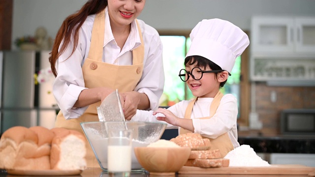 可爱的亚洲小男孩和美丽的母亲在家庭厨房的桌子上用筛网筛面粉准备烘焙和蛋糕。泰国小孩像厨师一样玩面粉视频素材