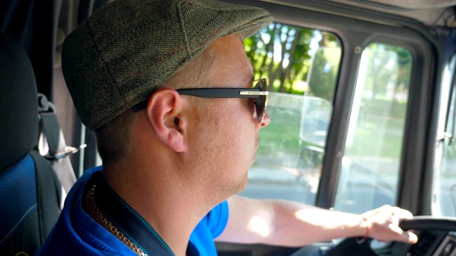 在阳光明媚的日子里，在城市道路上驾驶的卡车司机的侧面。戴着帽子和太阳镜的男人控制着他的卡车，专注地看着道路。内景慢镜头视频下载