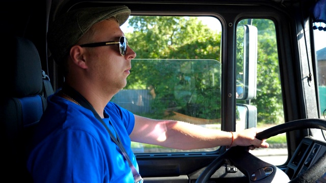 在阳光明媚的日子里，货车车厢内的司机在乡间小路上兜风。戴着帽子和太阳镜的男人控制着他的卡车，专注地看着道路。慢镜头视频下载