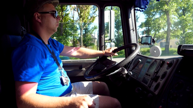 在阳光明媚的日子里，货车车厢内的司机在乡间小路上兜风。戴着帽子和太阳镜的男人控制着他的卡车，专注地看着道路。特写慢动作视频下载