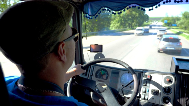 在阳光明媚的日子里，一名面目全非的卡车司机在县道上行驶。戴着帽子和太阳镜的男人控制着他的卡车，专注地看着道路。从船舱内看交通。内景慢动作特写视频下载