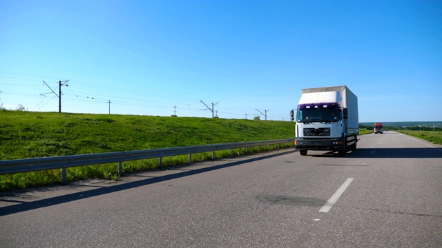 摄影机跟随卡车与货物拖车在公路上行驶在夏季的货物运输。白色的卡车驶过乡村，背景是美丽的风景。慢镜头前视图视频素材