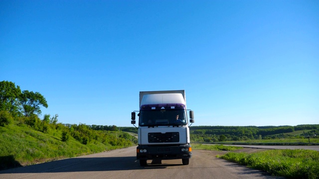 卡车正面视图与货物拖车行驶在公路上运输货物在夏季的一天。白色的卡车驶过乡村，背景是美丽的自然景观。慢动作视频素材