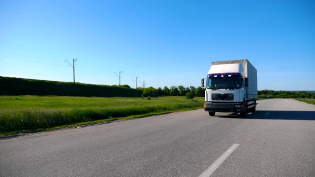 镜头跟随卡车与货物拖车在空旷的公路上行驶在夏季的货物运输。一辆白色卡车以自然景观为背景穿过乡村。慢镜头前视图视频素材