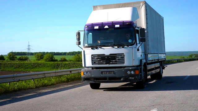 摄影机跟随卡车与货物拖车在公路上行驶在夏季的货物运输。白色的卡车驶过乡村，背景是美丽的风景。慢镜头前视图视频素材