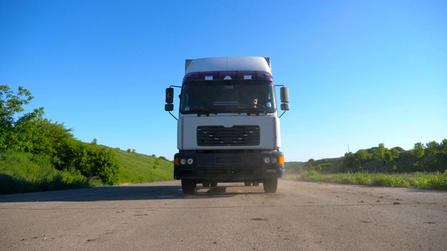 卡车正面视图与货物拖车行驶在公路上运输货物在夏季的一天。白色的卡车驶过乡村，背景是美丽的风景。慢镜头视频素材