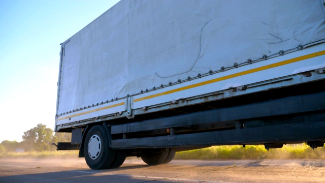 低视野的卡车与货物拖车行驶在高速公路和运输货物在日落时间。一辆白色卡车在乡间公路上疾驰，后面扬起尘土。物流的概念。慢动作视频下载