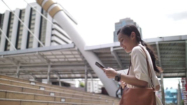 一名亚洲女性正在用智能手机和朋友在线聊天，走在办公大楼里视频素材