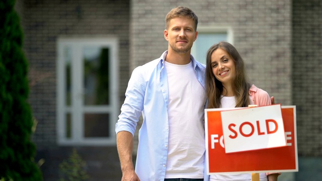 幸福的夫妇拿着房子的钥匙在出售标志附近，年轻家庭的抵押贷款视频下载