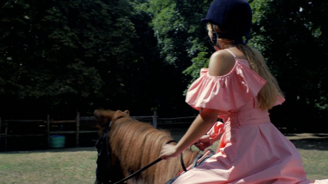 一个十几岁的女孩戴着保护头盔，穿着粉红色的衣服，骑着一匹棕色的马，背景是树木。侧视图。4 k。4 k的视频视频下载