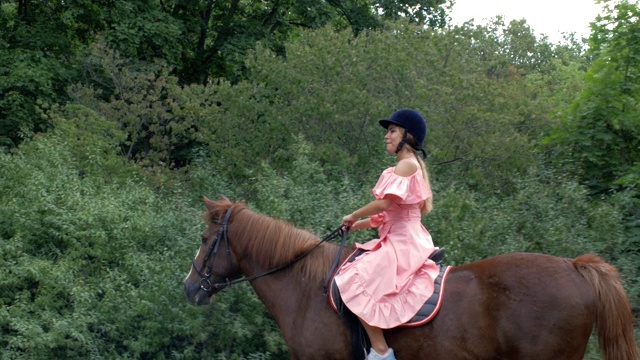 年轻的女孩戴着保护头盔，穿着粉红色的衣服，骑着一匹棕色的马，在树木和树叶的背景下。4 k。4 k的视频视频下载