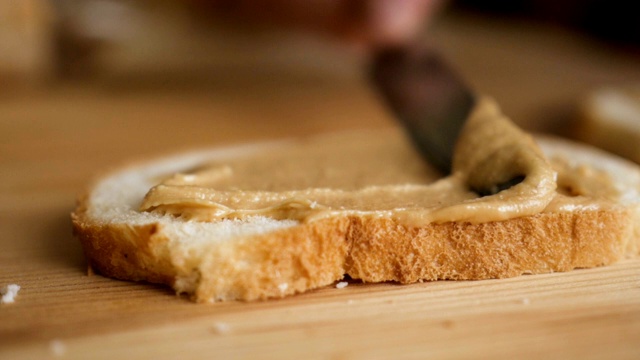 美味而丰盛的早餐，坚果奶油配脆面包。男人做了个花生酱三明治…男人的手在厨房的面包上涂抹黄油的特写，慢镜头。视频素材