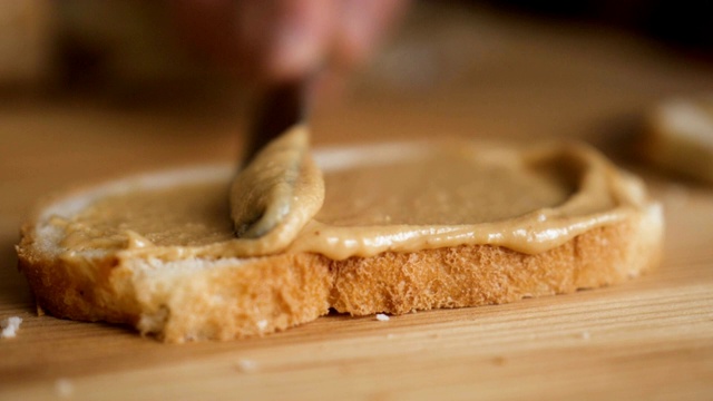 美味而丰盛的早餐，坚果奶油配脆面包。男人做了个花生酱三明治…男人的手在厨房的面包上涂抹黄油的特写，慢镜头。视频素材