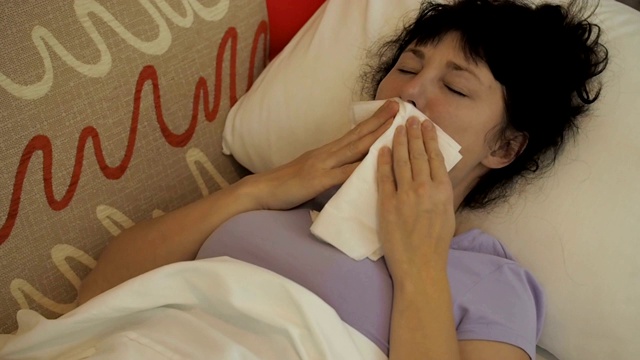 一个生病的女人坐在家里的沙发上打喷嚏。女孩流鼻涕，拿纸巾，流感症状。医疗保健和医疗理念。感冒是流感的感染。视频素材