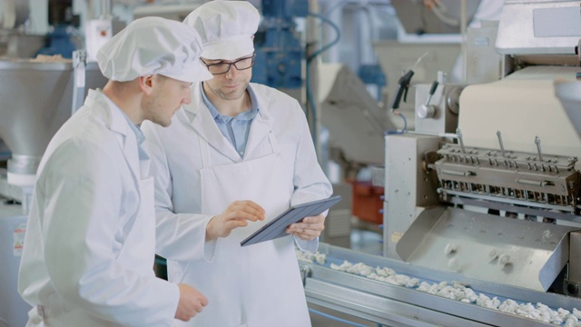 两名年轻男性质量主管或食品技术人员正在检查饺子食品厂的自动化生产。7 .员工使用平板电脑工作。他们穿着白色的工作袍。视频素材