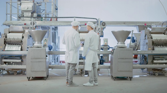 两个年轻的食品工厂员工讨论与工作有关的事情。男性技术员或质量经理使用平板电脑工作。他们戴着白色的卫生帽和工作袍。视频素材