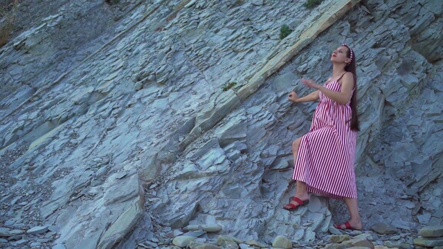 年轻的怀孕女孩穿着红白相间的条纹连衣裙。一个女孩站在一座岩石山的脚下，抚摸着自己的肚子。视频素材