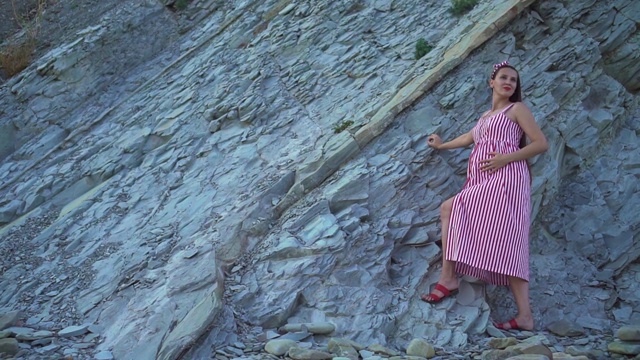 年轻的怀孕女孩穿着红白相间的条纹连衣裙。一个女孩站在一座岩石山的脚下，抚摸着自己的肚子。视频素材
