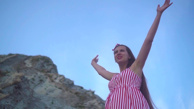 一个年轻的怀孕女孩在一个岩石山脚下穿着红色和白色条纹的裙子，举起手来表示自由。视频素材
