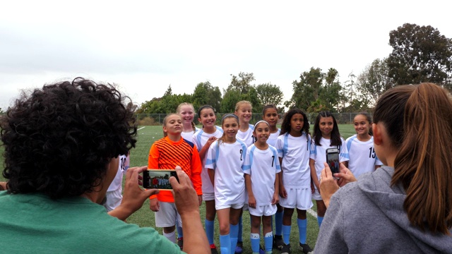赛后，父母们用智能手机拍摄年轻女子足球队在赛场上的照片视频素材