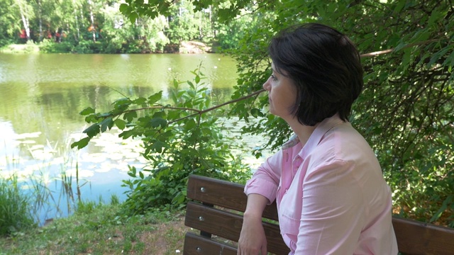 一位中年妇女坐在池塘边的长凳上休息。视频下载