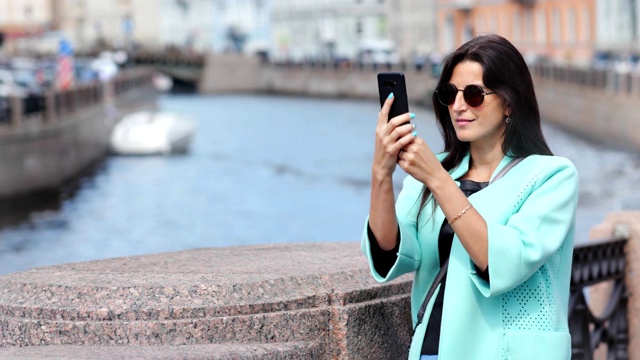 微笑的时尚女游客站在河上的桥上用智能手机拍照视频素材