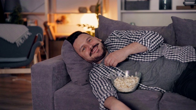 一个快乐的人在晚上在家微笑着看电视吃爆米花的肖像视频素材