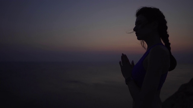 平静和女孩练习瑜伽和冥想日落在海边，在悬崖上。女运动员在祈祷时手拉手。精神性，正念和健康生活方式的概念。视频素材