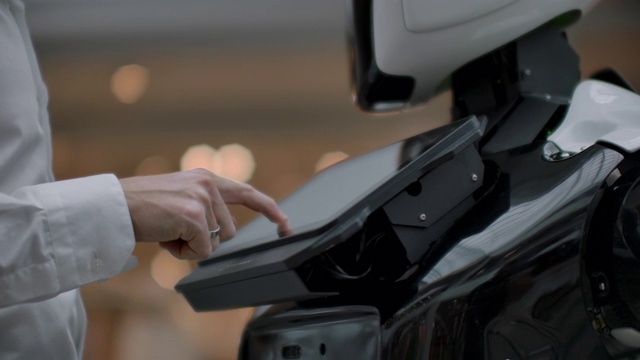 一名身穿衬衫的男子与一个白色机器人交流，机器人问问题，并用手指按屏幕。视频素材
