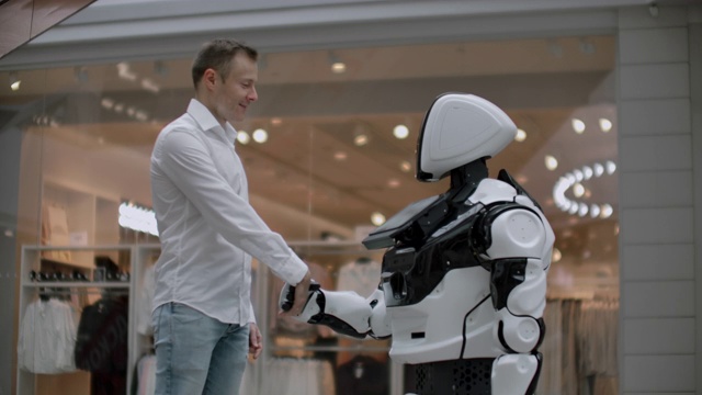 一名身穿衬衫的男子与一个白色机器人交流，机器人问问题，并用手指按屏幕。视频素材