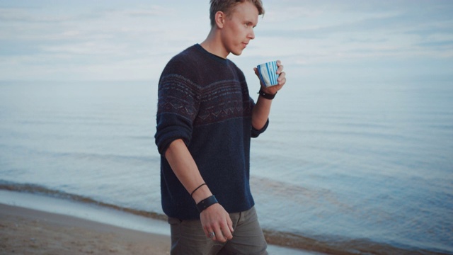 年轻英俊的男人在海滩上散步。平静的大海在一个多云温暖的早晨。他正在用茶杯喝水。他穿着正宗的针织毛衣和短裤。海边浪漫的夏日时光。视频下载