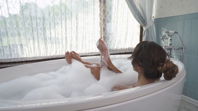 一个年轻迷人的女人有泡沫浴和看窗户在时尚的室内浴室的后视图。旋转镜头视频素材