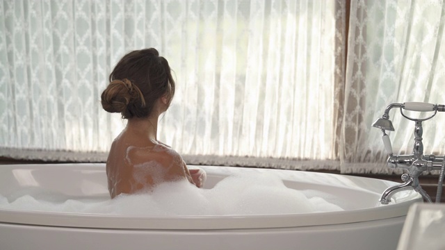 一个年轻迷人的女人与美丽的金色日光浴泡泡浴和看窗户在时尚的室内浴室。视频下载
