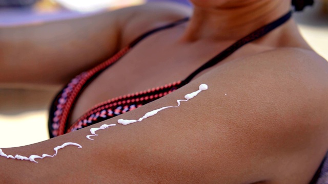 女模特在手上和肩膀上涂防晒霜，坐在沙滩上，像电影一样视频素材