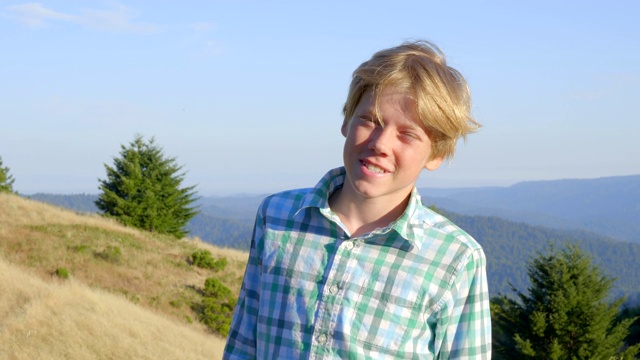 一个漂亮的金发男孩的肖像，12岁，看着相机的慢动作。积极的情感。美丽的风景背景。日落时间。视频下载