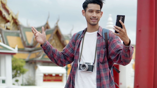 年轻英俊的亚洲男子独自站在街上用智能手机与朋友视频聊天，背景是泰国的寺庙。快乐享受休闲旅游生活方式。视频素材