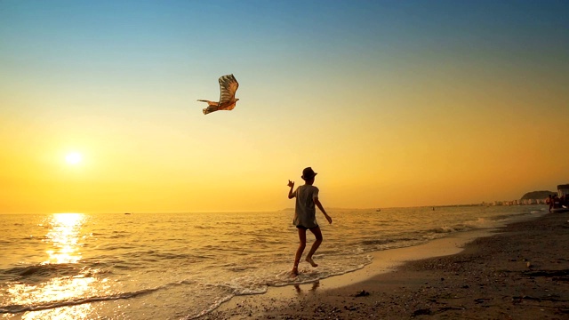 一个小男孩放着风筝在空旷的海滩上奔跑，背景是令人惊叹的夏日日落视频素材